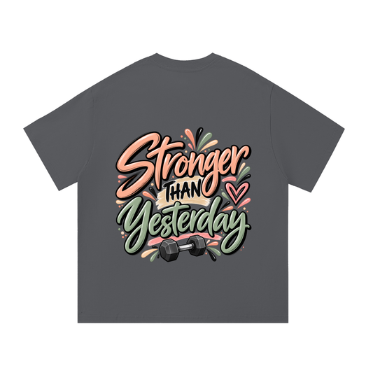 A dark gray t-shirt with a colorful graphic on the front that reads 'Stronger Than Yesterday' in stylized peach, black, and green lettering, decorated with a heart and a dumbbell.